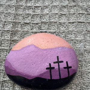 May include: A smooth, rounded river stone painted with a landscape scene. The design features a black foreground with three black crosses, a purple mid-ground representing hills, and a light peach-colored sky at the top of the stone.