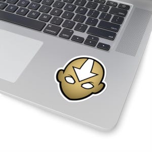 Könnte beinhalten: Ein Aufkleber mit einer stilisierten Darstellung eines Kopfes mit goldfarbenem Gesicht, weißen Augen und einem weißen Pfeil auf der Stirn. Der Aufkleber befindet sich auf einem silbernen Laptop.