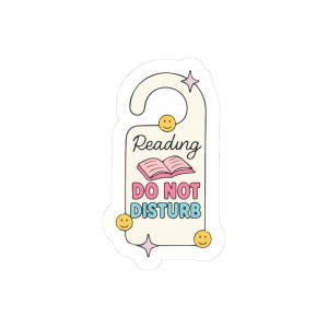 May include: A cream-coloured door hanger sticker with the words "Reading DO NOT DISTURB" in a retro font. The sticker features a pink open book graphic, smiley faces, and star accents. Ideal for book lovers.