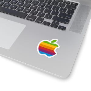 Puede incluir: Una pegatina colorida con el icónico logotipo de Apple con un diseño de arcoíris. La pegatina está colocada en un portátil plateado, mostrando una vibrante mezcla de colores que incluyen rojo, naranja, amarillo, verde, azul y morado. La pegatina tiene un borde blanco.