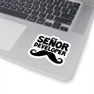 May include: A white sticker with the text "SEÑOR DEVELOPER" in black, featuring a black mustache graphic. The sticker is placed on a silver laptop, with the keyboard visible.