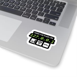 May include: A white sticker featuring the text "THERE'S NO PLACE LIKE 127.0.0.1" in black and green. Below the text are icons of a computer, Wi-Fi, and a bed. The sticker is placed on a laptop.