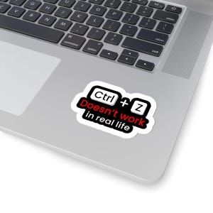Può includere: Un adesivo bianco con la scritta "Ctrl + Z Doesn't work in real life" in nero e rosso. L'adesivo è su un laptop argentato, vicino alla tastiera.