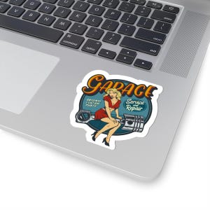 May include: A sticker featuring a vintage garage theme. The design includes the word "GARAGE" in a stylized font, a woman in a red dress, and the text "Service and Repair" and "Original Custom Parts". The sticker is on a laptop.