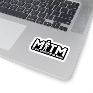May include: A white sticker with a black border and the text "MTM YOUR SESSION IS MINE" in a bold, sans-serif font. The sticker is placed on a silver laptop, with the keyboard visible in the background.
