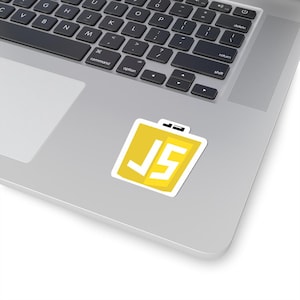 May include: A yellow sticker with a white border featuring the letters "JS" in a bold, white font. The sticker is placed on a silver laptop, with the keyboard visible. The sticker has a small black rectangle at the top.