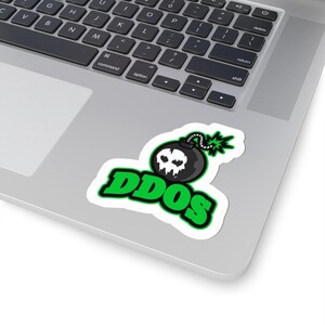 Può includere: Un adesivo bianco con una bomba nera con un teschio e dettagli verdi. La parola "DDOS" è scritta in grassetto verde sotto la bomba. L'adesivo è posizionato su un laptop.