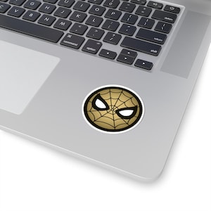 May include: A circular sticker with a gold spiderweb design, white eye shapes, and a black border. The sticker is on a silver laptop.