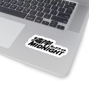Op de afbeelding: Een witte sticker met het woord "MIDNIGHT" in een vetgedrukte, zwarte, gestileerde letter. Japanse karakters staan erboven. De sticker bevindt zich op een zilveren laptop.