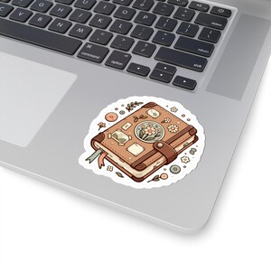 May include: A sticker featuring a brown, illustrated journal with a leather strap and floral accents. The journal has a ribbon bookmark and is surrounded by small floral and leaf illustrations. The sticker is on a laptop.