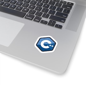 May include: A white-bordered sticker featuring the C++ programming language logo, a white "C" with two plus signs, set against a blue and white hexagonal background. The sticker is placed on a laptop.