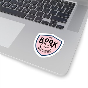 Può includere: Un adesivo con un design a scudo rosa, con la faccia di un gatto e la scritta "THE BOOK WAS BETTER" in nero. L'adesivo ha un bordo bianco e un contorno blu, posto su un laptop.