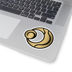 Könnte beinhalten: Ein gold-schwarzer Aufkleber mit weißem Rand. Der Aufkleber zeigt ein stilisiertes Design aus Mond und Kreis. Der Aufkleber befindet sich auf einem silbernen Laptop.
