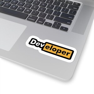 May include: A white sticker with the word "Developer" in black, outlined in white, and set against a yellow rectangle. The sticker is placed on a silver laptop, with the keyboard visible.