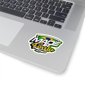 May include: A die-cut sticker featuring the text MADE IN FAVELA in bold white and yellow lettering with a black outline. The design incorporates a stylized green, yellow, and blue Brazilian flag element and an Instagram camera icon.
