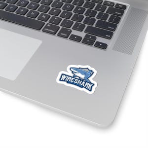 May include: A white sticker with a blue shark graphic and the text "WIRESHARK NETWORK SNIFFER" in blue and white. The sticker is placed on a laptop.