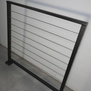 May include: Black metal railing with horizontal stainless steel cables. The railing has a rectangular frame and is mounted on two square posts. The design is modern and minimalist, suitable for decks or balconies.