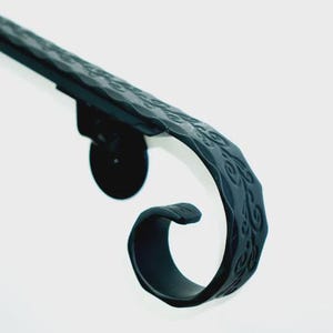 May include: Close-up of a black, ornate metal handrail. The handrail features a textured surface with a repeating pattern and a curved, scroll-like end. The design suggests a decorative element for interior or exterior use.