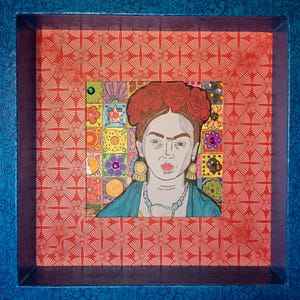 May include: Framed artwork depicting Frida Kahlo. The portrait is set against a red patterned backdrop within a blue frame. The artwork incorporates a colourful mosaic and is embellished with decorative elements.