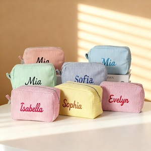 Puede incluir: Una colección de estuches rectangulares con cremallera hechos de tela a rayas en varios colores pastel, incluyendo rosa, azul claro, verde menta, amarillo y azul marino. Cada estuche presenta un nombre bordado en letra cursiva.