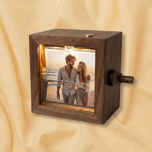 May include: A square dark wood frame containing a backlit photograph of two people standing on a beach. The frame features a small brass-colored button on the top edge and a wooden knob on the right side for rotating the internal display mechanism.