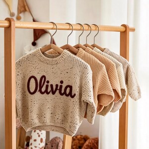 Op de afbeelding: Een rij kindertruien in verschillende neutrale kleuren hangt aan een houten rek. De eerste trui is beige met donkere spikkels en de naam "Olivia" in een donkerbruin script. De andere truien zijn effen in tinten van bruin en crème.