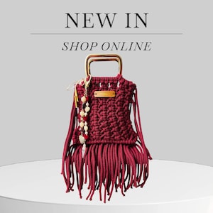 May include: A burgundy macrame handbag with a gold-colored handle and fringe detail. The bag has a woven design and a small wooden tag. The words "NEW IN" and "SHOP ONLINE" are displayed above the bag.