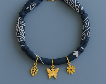 May include: A navy blue fabric necklace with a white paisley pattern. The necklace features three gold-colored charms: a leaf, a butterfly, and a flower. The necklace is connected with a gold chain and is displayed on a blue background.