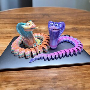 Könnte beinhalten: Zwei farbenfrohe Kobra-Figuren. Eine ist hauptsächlich blau und orange, die andere lila und rosa. Beide haben einen gewundenen Körper und eine erhobene Haube mit cartoonartigen Gesichtszügen. Sie liegen auf einer dunkelgrauen Oberfläche.