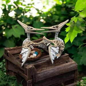 Op de afbeelding: Twee Pterodactylus skeletfiguren met uitgestrekte vleugels, geplaatst op een verweerde houten kist. Een klein nest met blauwe en witte eieren staat ertussen. De achtergrond toont weelderig groen gebladerte.