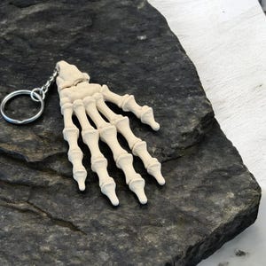 Op de afbeelding: Een beige sleutelhanger in de vorm van een skelet hand met een zilveren sleutelring. De hand is gedetailleerd met individuele vingerkootjes. De sleutelhanger ligt op een donkergrijze, getextureerde ondergrond. Uniek accessoire.