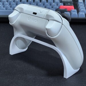May include: A white Xbox controller on a white stand. The controller features the Xbox logo and a black joystick. The stand has a curved design, supporting the controller.
