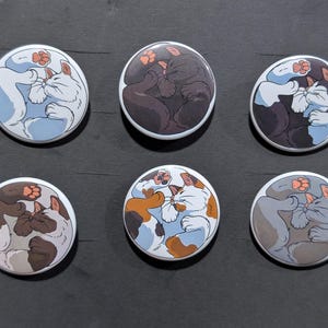 May include: A set of six circular pinback buttons, each featuring a different illustration of a curled-up sleeping cat. The cats are depicted in various colors including white, black, brown, calico, and gray, all showing visible paw pads.