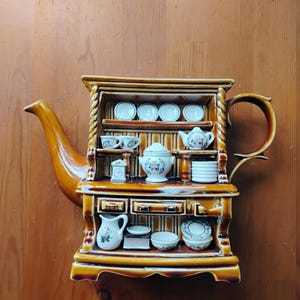 May include: A ceramic teapot shaped like a miniature hutch cabinet with a glossy brown finish. The front features open shelves displaying tiny white porcelain dishes, including plates, cups, a lidded tureen, a pitcher, and a small teapot.