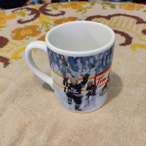 May include: A white ceramic mug featuring a colourful graphic of two young hockey players on the ice. The players wear dark blue jerseys with the text Tim Hortons printed on the front. The background shows a hockey net and a crowd of spectators.