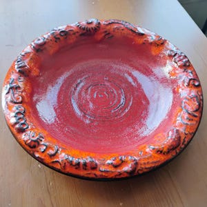 May include: A round ceramic bowl with a glossy finish, featuring a vibrant red centre with concentric circular indentations. The wide, raised rim is textured with dark, swirling patterns and shades of bright orange and deep red throughout.