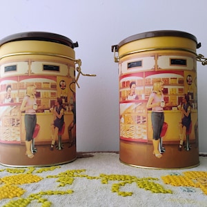 May include: Two cylindrical metal canisters with brown lids and gold-coloured clasps. The canisters feature a vintage diner scene with women and shelves of food. The background is a light yellow colour.