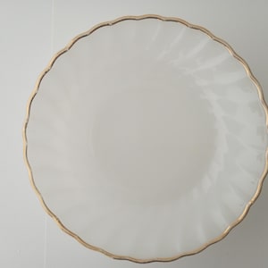 May include: A white plate with a gold rim. The plate has a scalloped edge and a subtle swirl pattern. The gold rim adds a touch of elegance to the simple design.