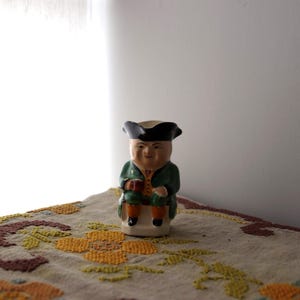 Könnte beinhalten: Keramikfigur eines sitzenden Mannes in einem grünen Mantel, schwarzem Hut und orangefarbener Kniehose. Die Figur hält einen kleinen roten Gegenstand. Die Figur steht auf einem weißen Sockel.