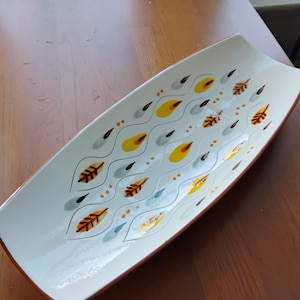 May include: A rectangular ceramic serving tray with a white base featuring a repeating pattern of yellow, grey, and brown leaf-like shapes arranged within thin, wavy black lines, accented by small brown dots throughout the design.