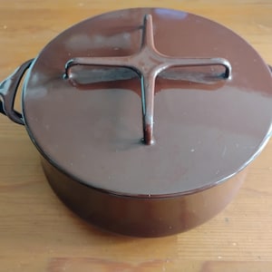 Puede incluir: Una olla redonda de hierro fundido esmaltado en color marrón oscuro con tapa a juego. La tapa presenta un distintivo mango en forma de cruz centrado en la parte superior. Dos asas curvas están integradas en los lados de la olla.