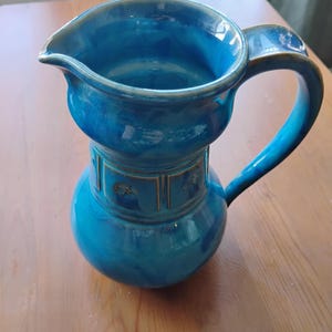 May include: A ceramic pitcher with a glossy, vibrant blue glaze featuring a rounded body, a wide pouring spout, and a curved handle. The midsection of the pitcher is decorated with a series of rectangular panels containing embossed circular motifs.