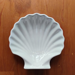 Puede incluir: Un plato de cerámica blanca con forma de concha de vieira. El plato presenta un acabado brillante y crestas radiantes distintivas que se extienden desde la base hasta el borde festoneado, creando una superficie cóncava que imita la forma natural.