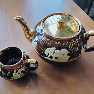Puede incluir: Una tetera y una jarra de leche de cerámica marrón oscuro con un acabado dorado reflectante en el pico, el asa, la tapa y el borde. Las piezas están decoradas con figuras en relieve de color crema y líneas verticales doradas.