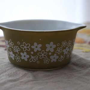 Op de afbeelding: Een olijfgroene Pyrex-schaal met witte bloemmotieven. De schaal heeft een wit interieur en twee kleine handvatten. De schaal staat op een beige en patroon oppervlak. Een vintage keukenartikel.