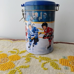 May include: A cylindrical tin container with a blue lid and a metal clasp. The container features a hockey scene with players in blue and red jerseys. The text "Tim Hortons" is visible on the blue jersey. The background shows a crowd of people.