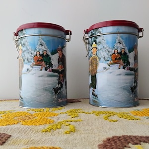 May include: Two cylindrical metal canisters with red lids and a winter scene illustration. The illustration depicts people ice skating and sitting on a bench near snow-covered trees. The canisters have a hinged closure.