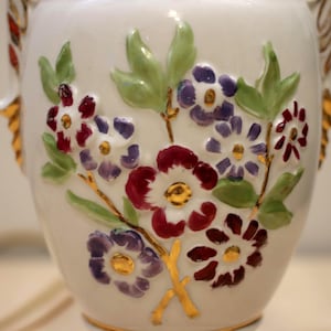 May include: A white ceramic vase with a floral design. The vase features raised relief flowers in shades of purple, red, and white, with green leaves and gold accents. The vase has two handles.