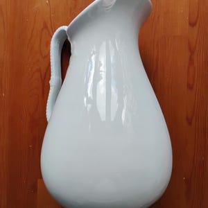 Puede incluir: Una jarra de cerámica blanca brillante de gran tamaño con cuerpo redondeado y asa curva que presenta un diseño texturizado de cuentas en el borde exterior. La jarra tiene un pico vertedor ancho en la parte superior con una pequeña muesca.