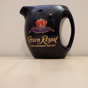 Può includere: Una brocca in ceramica blu navy con un manico grande e un beccuccio. La brocca presenta un logo dorato con una corona e le parole "Crown Royal" e "The Legendary Import."